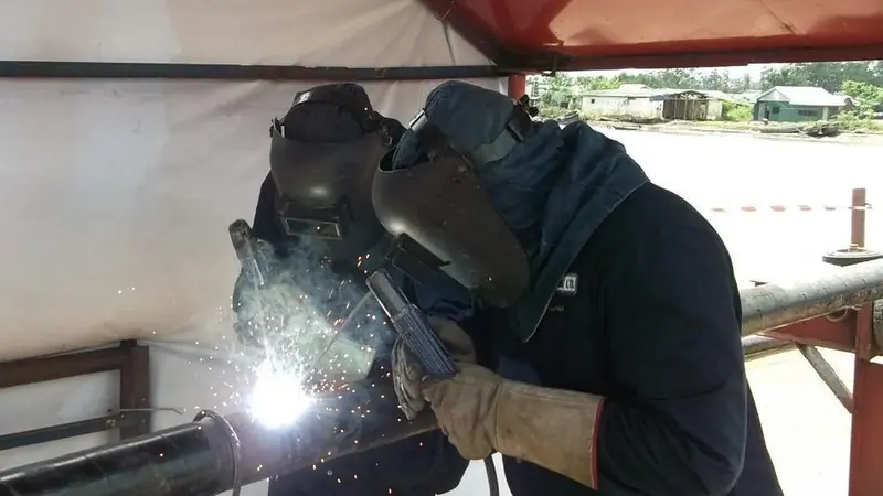 Certified welder completing a pipeline tie-in weld with a second hand holding the shield